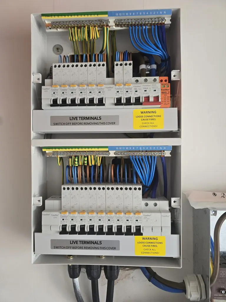 Open electrical consumer unit featuring two rows of circuit breakers with labeled warning stickers about live terminals and loose connections.