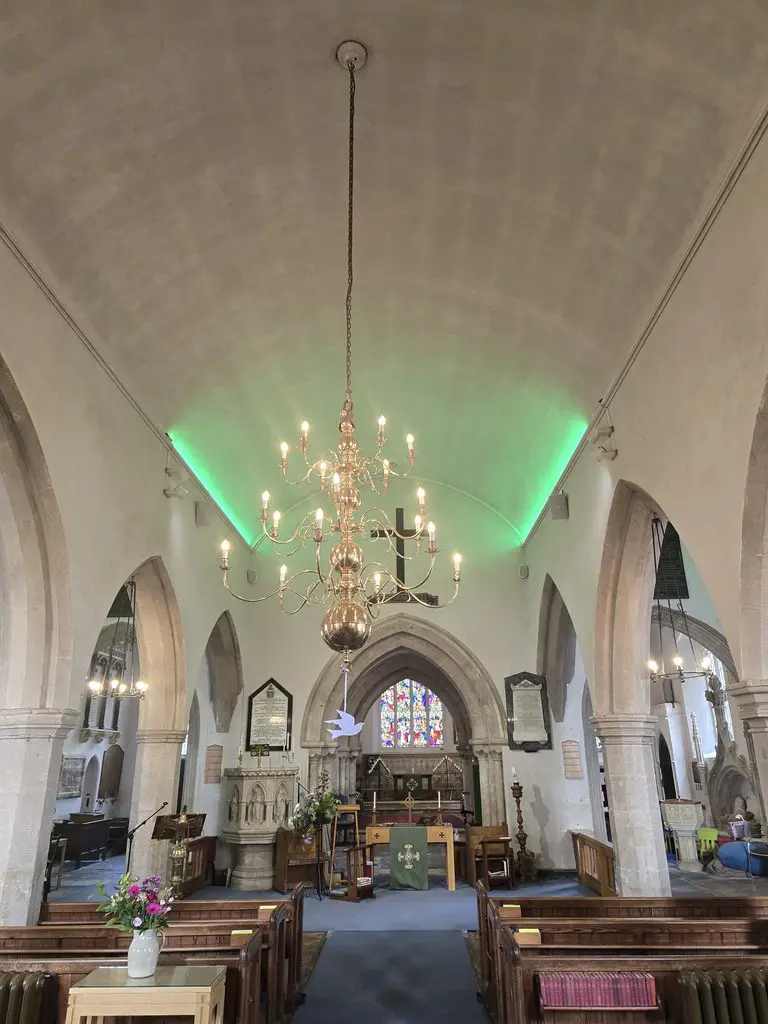 Interior of a stone church featuring a large brass chandelier, wooden pews, a central altar, and a green light shining along the vaulted ceiling.