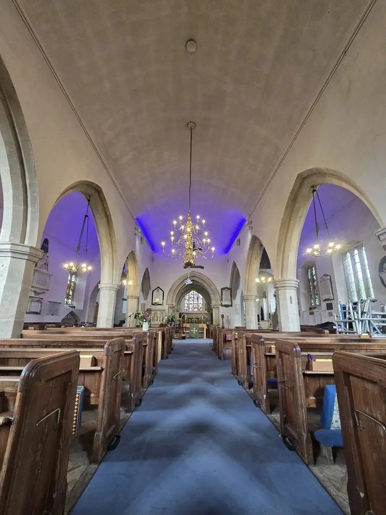 Interior view of a church looking down the central aisle toward the altar, featuring wooden pews, stone arches, chandeliers, and purple accent lighting along the ceiling.
