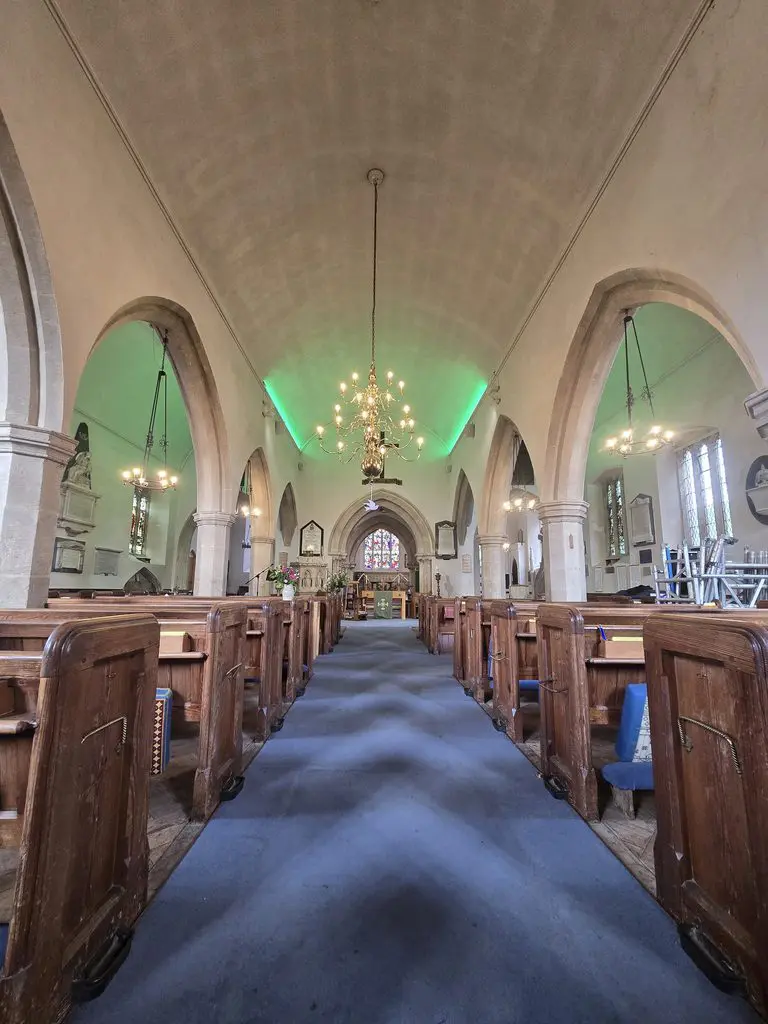 Interior view of a church nave with wooden pews lining a blue carpeted center aisle, featuring stone arches and a green-lit arched ceiling leading toward the altar.