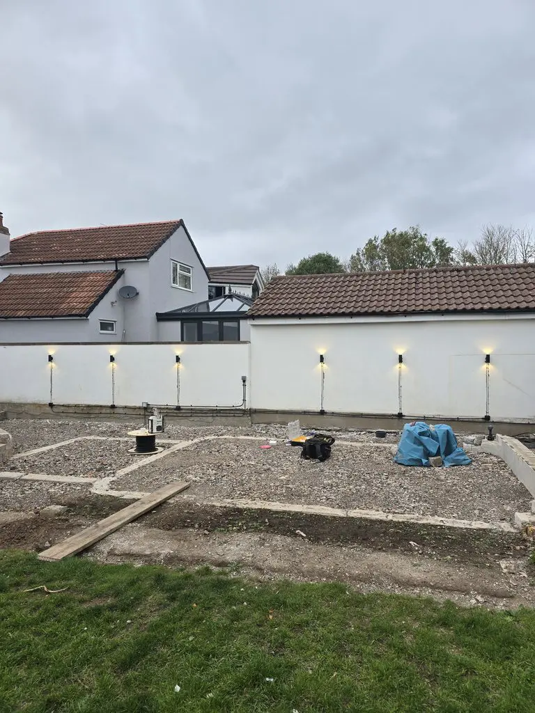 A gravel-covered backyard under construction, featuring a white wall with installed outdoor lights and various tools scattered on the ground.