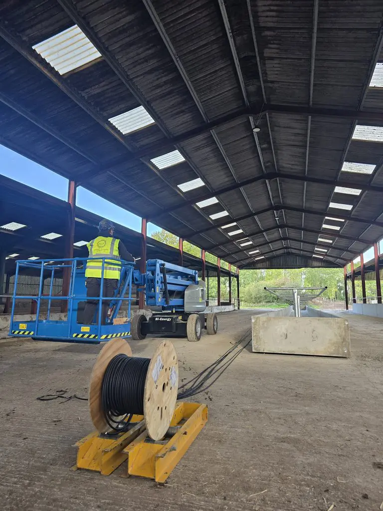 A construction worker in a high-visibility vest operates a blue scissor lift inside a large open-sided farm building, with a spool of black cable in the foreground.