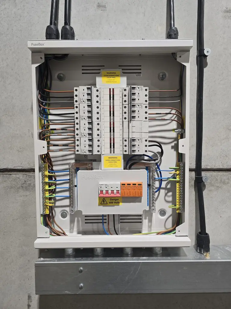An open FuseBox electrical distribution board mounted on a concrete wall, showing circuit breakers, wiring, and safety warning labels.