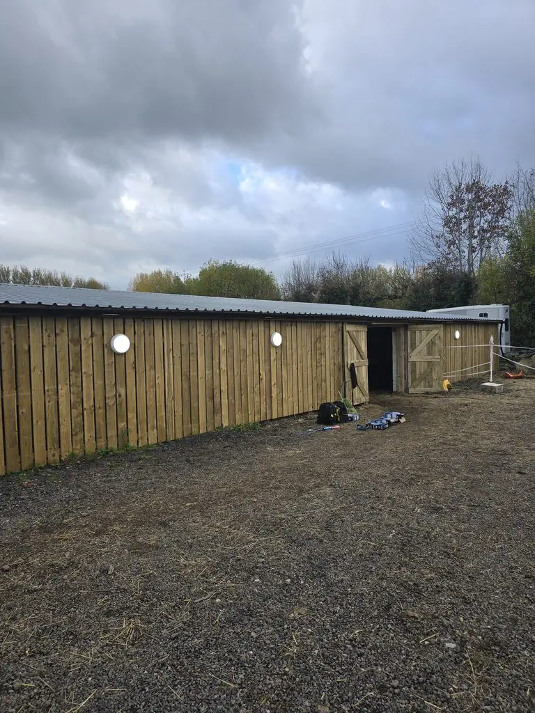 A long wooden stable building with a metal roof and open double doors on a gravel lot under a cloudy sky.