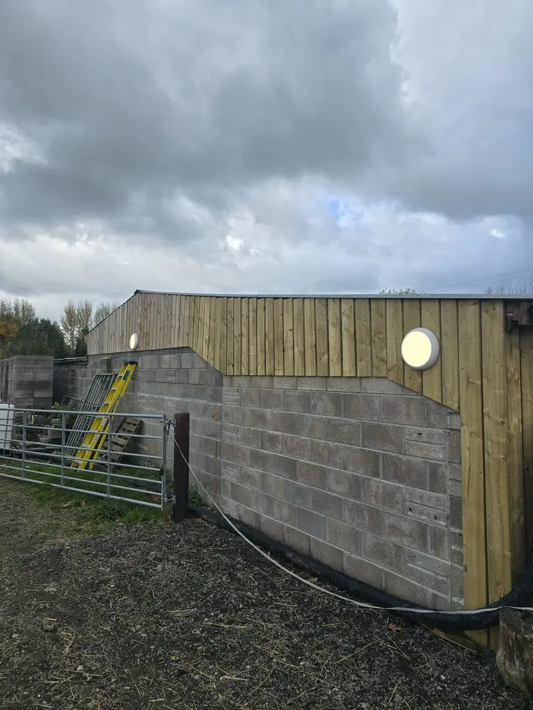 A partially wood-clad cinder block wall with two circular outdoor lights, a yellow ladder leaning against the side, and a metal gate in the foreground under a cloudy sky.