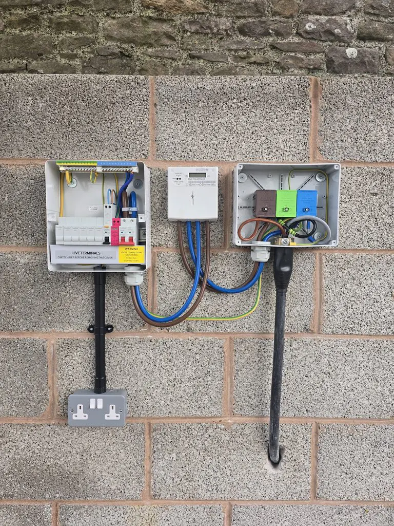 Electrical components mounted on a stone wall, including a consumer unit, an electricity meter, and a terminal block housing, connected by various colored cables.