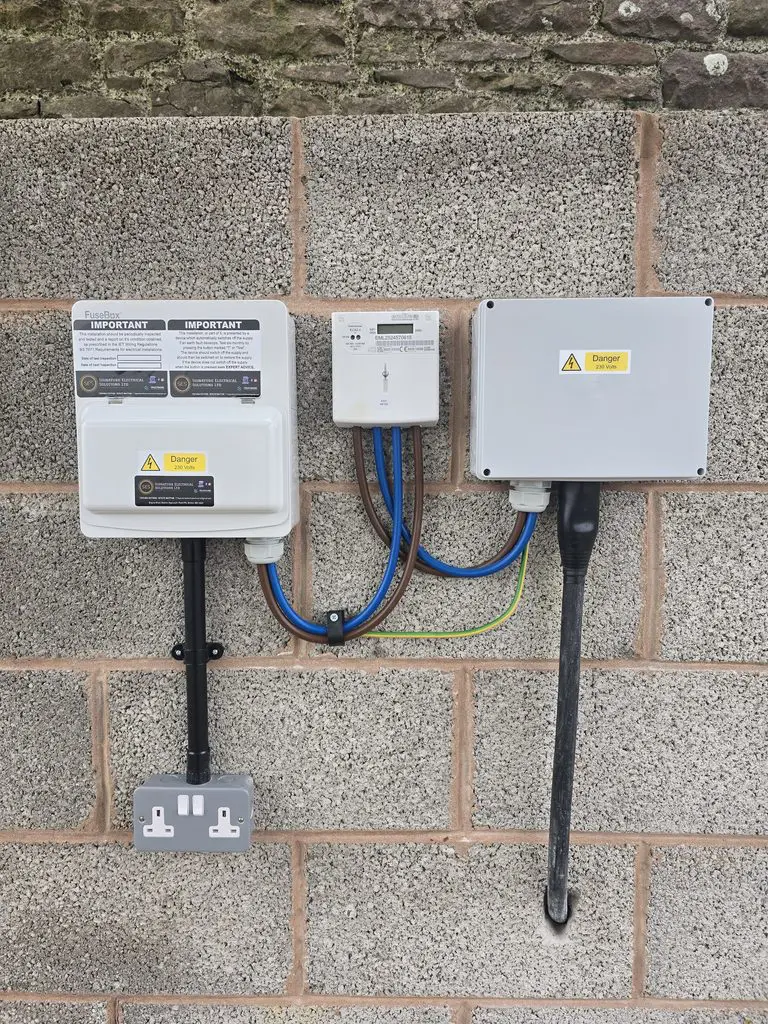 An electrical setup mounted on a stone wall, featuring a consumer unit, an electricity meter, and a junction box with connected wiring and a double power outlet.