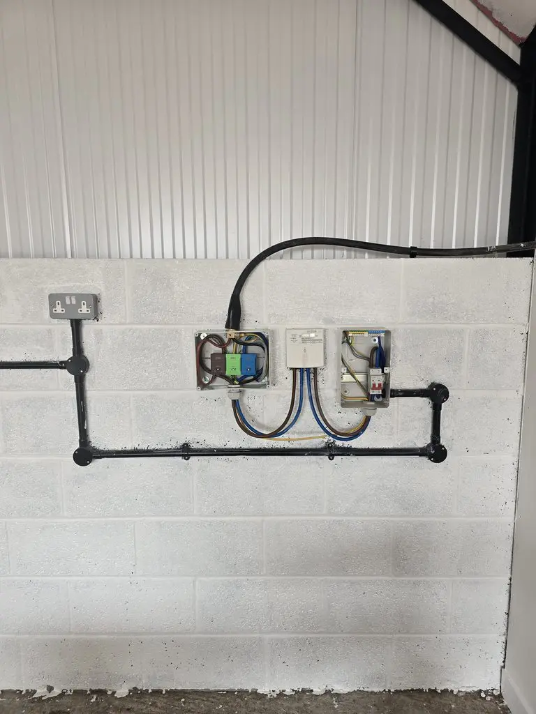 Electrical wiring and components mounted on a white cinder block wall, including a dual power outlet, two open junction boxes with visible wiring, and black conduit piping.
