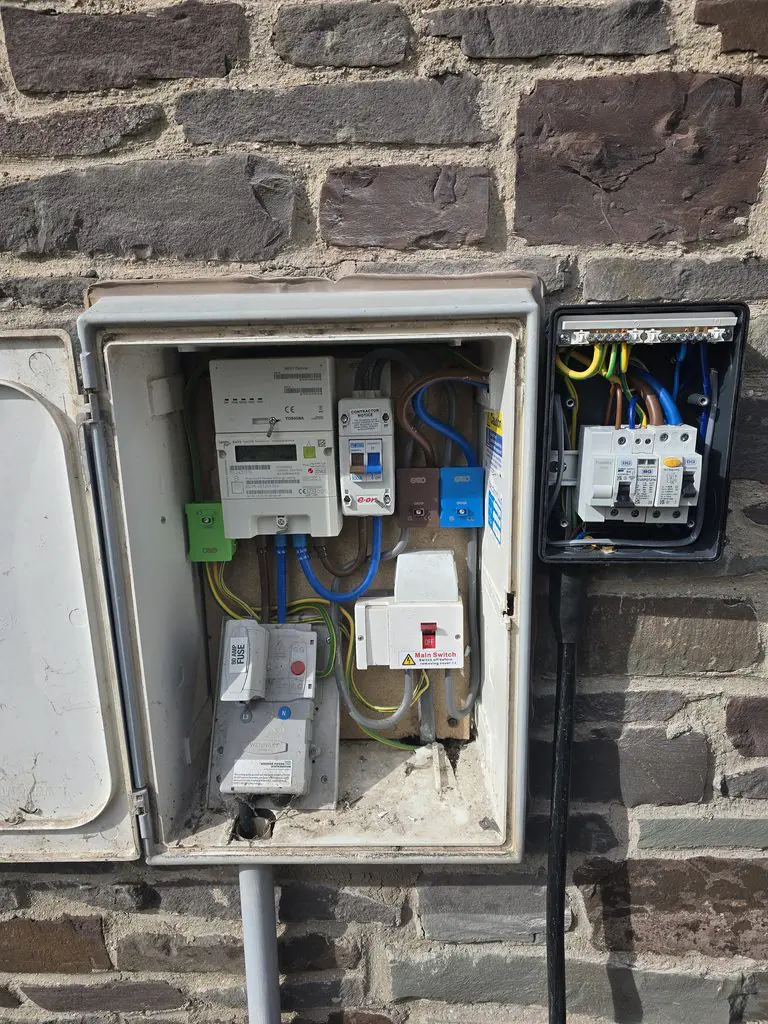 Open electrical meter boxes mounted on a stone wall, showing various breakers, a main switch, a fuse, and exposed wiring.