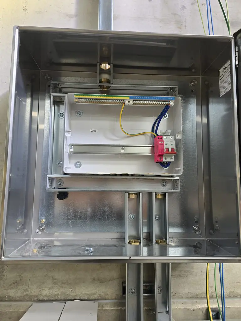 Open stainless steel electrical enclosure containing a white distribution box with a terminal block and a single red circuit breaker.