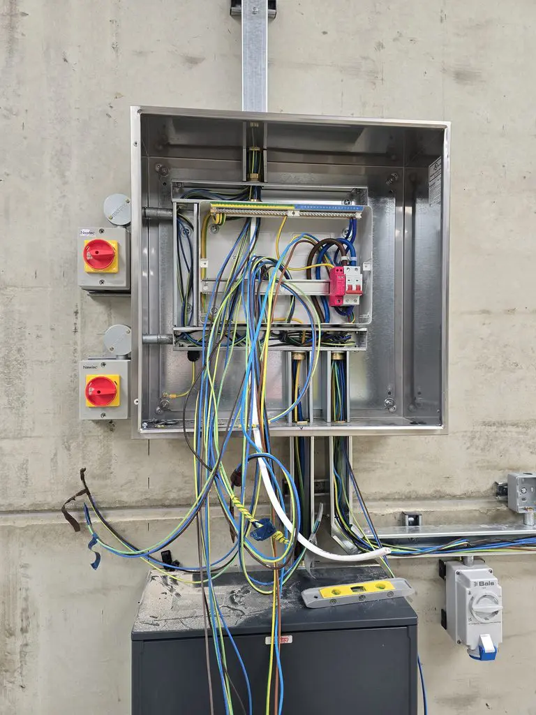 An open electrical junction box mounted on a concrete wall, featuring internal wiring, a terminal block, and two red rotary switches on the left side.