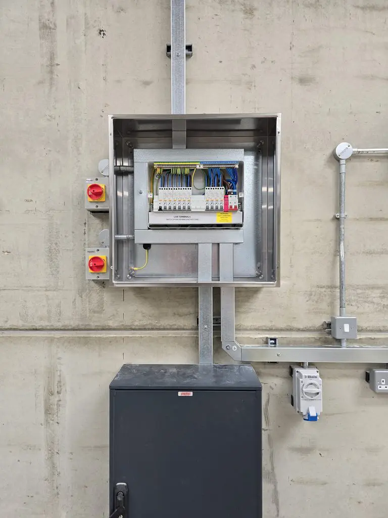 An open electrical control panel mounted on a concrete wall, featuring internal wiring, terminal blocks, two external red rotary switches, and a grey floor-standing cabinet below.