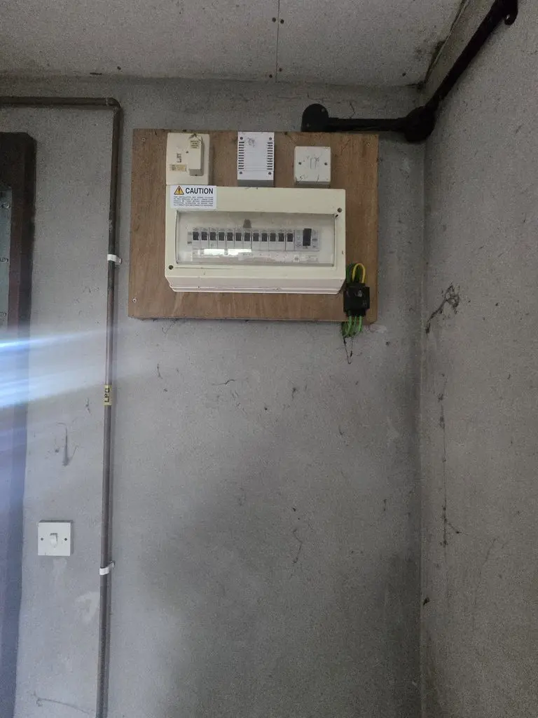 An electrical consumer unit mounted on a wooden board against a concrete wall, with an adjacent copper pipe labeled “LPG.”