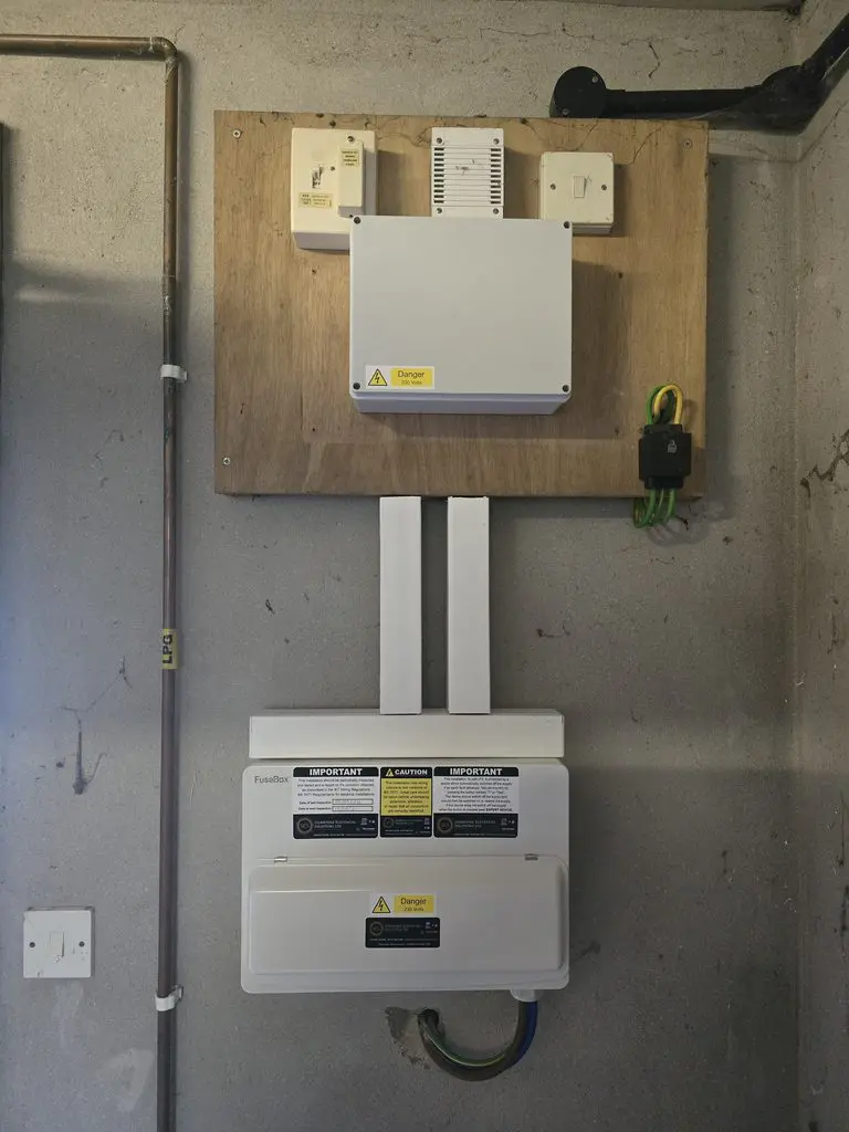An electrical consumer unit and a white distribution box mounted on a wooden board against a concrete wall.