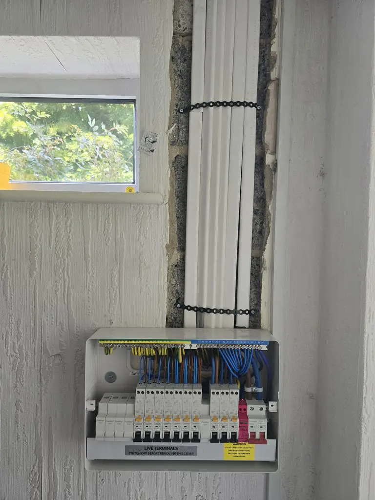 An open consumer unit with wiring, circuit breakers, and a “Live Terminals” warning label, mounted on a wall with white electrical conduits running vertically above it.