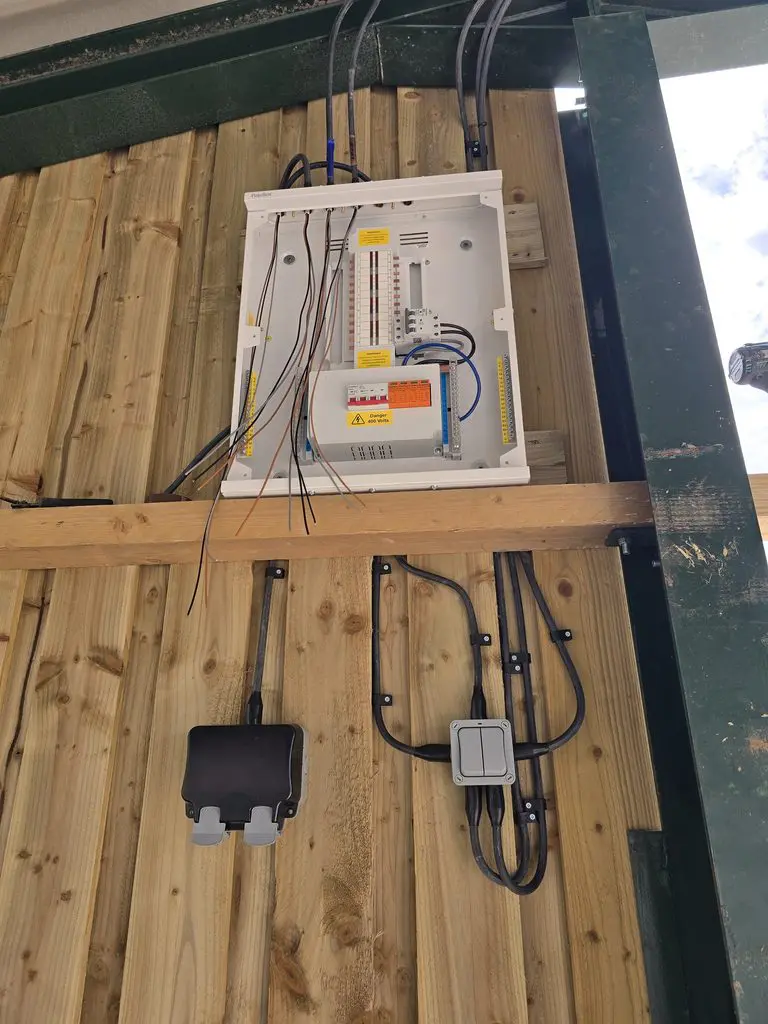 Electrical consumer unit mounted on a wooden wall, with wiring hanging from the top and two electrical boxes below.
