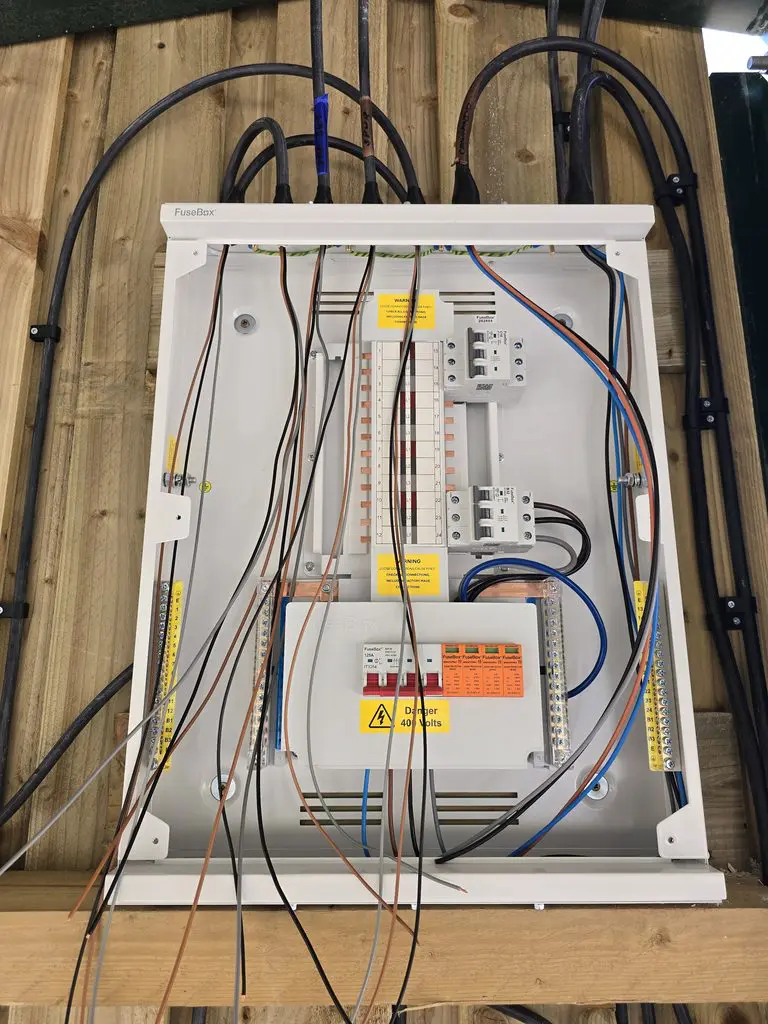 An open electrical consumer unit mounted on a wooden wall with various cables connected to circuit breakers and terminal blocks.