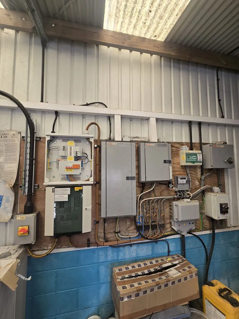 An array of electrical breaker boxes, control panels, and wiring mounted on a wooden board against a corrugated metal wall.