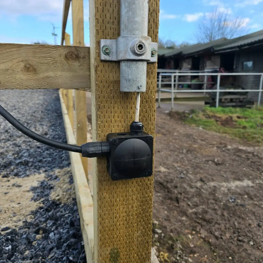 A black electrical junction box mounted to a wooden fence post, with cables running into its sides.