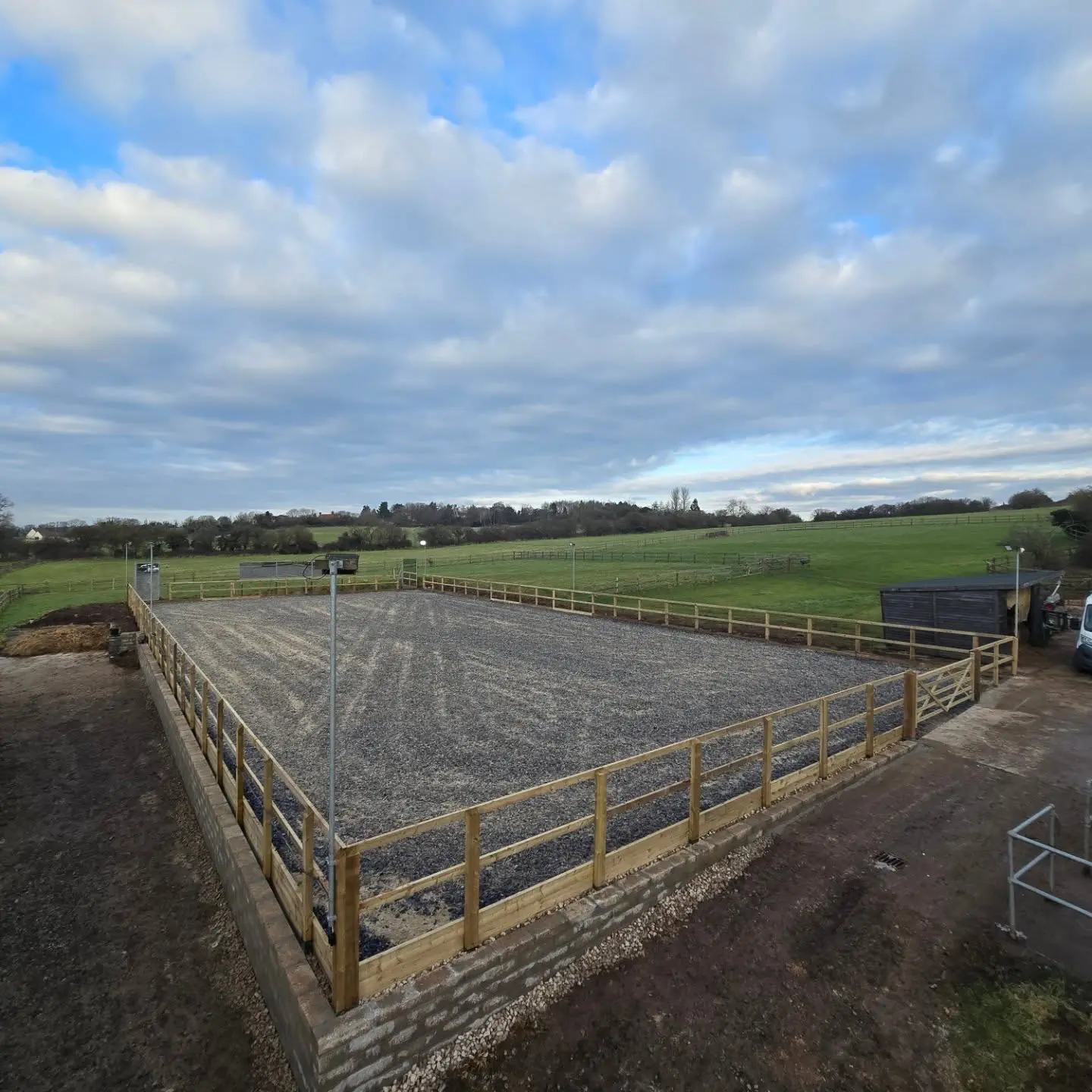 An outdoor horse riding arena with a gravel surface enclosed by a wooden fence, set in a grassy field under a cloudy sky.