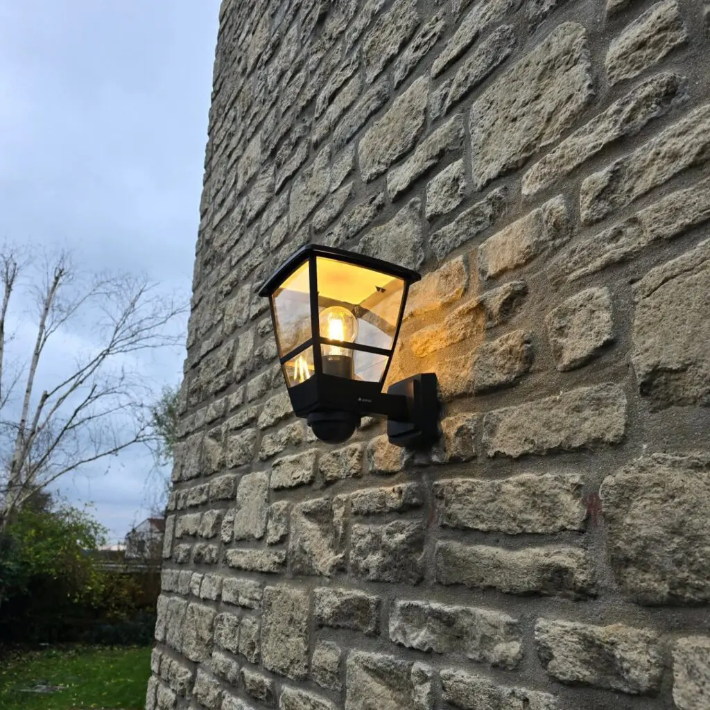 A black outdoor lantern-style wall light mounted on a stone wall, glowing with a warm yellow light.