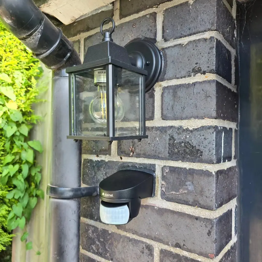 A black outdoor lantern fixture and a Steinel motion sensor mounted on a dark brick wall next to a downspout.