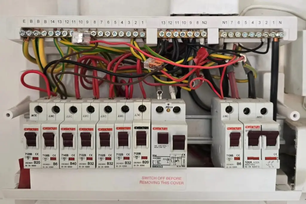 An open electrical consumer unit with circuit breakers, wiring, and terminal blocks, featuring a warning label that reads “SWITCH OFF BEFORE REMOVING THIS COVER.”