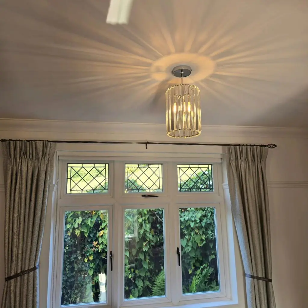 A cylindrical crystal pendant light casting a starburst pattern on the ceiling above a window with leaded glass panes and floor-length curtains.