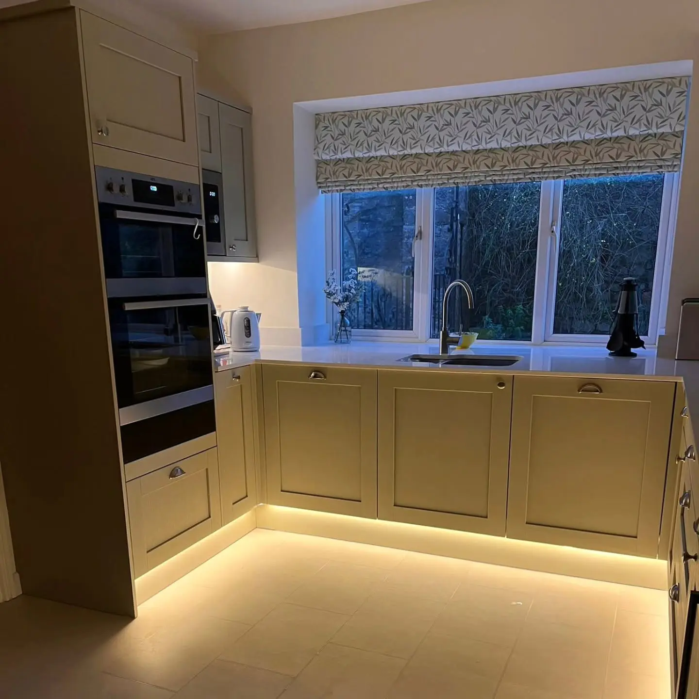 A kitchen corner with light-colored cabinets, a built-in wall oven, a sink under a window with a patterned roman shade, and under-cabinet LED lighting.