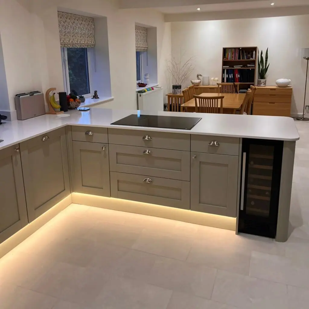 An L-shaped kitchen island with light gray cabinets, white countertops, a built-in induction cooktop, and an integrated wine cooler, featuring under-cabinet lighting.
