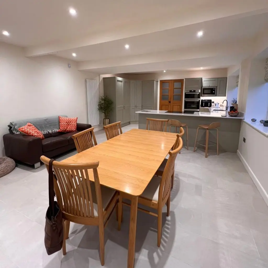 A wooden dining table with six chairs, positioned in an open-plan room with a kitchen island and a dark leather sofa.