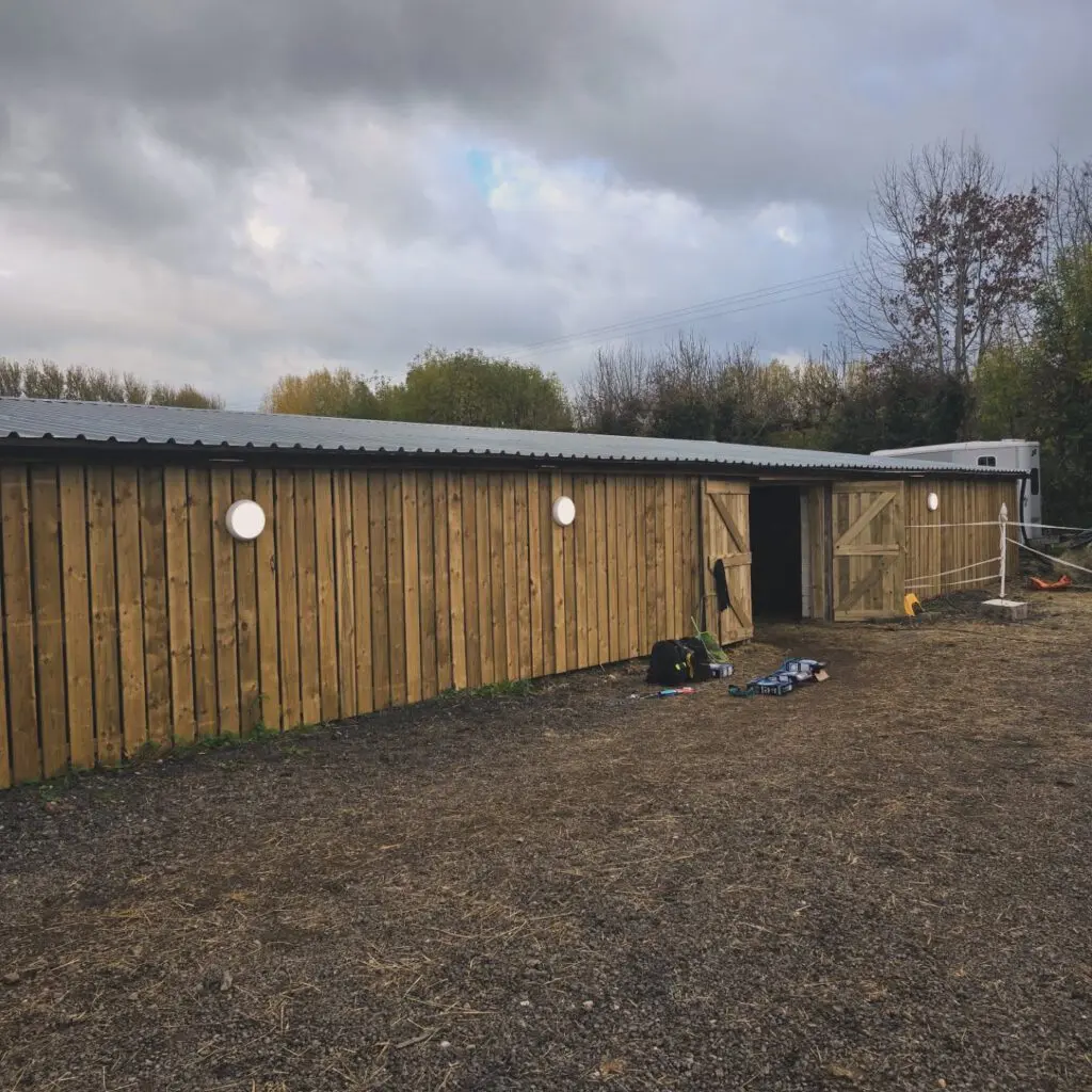 Long, single-story wooden building with a corrugated metal roof and round outdoor lights, featuring partially open double doors and construction tools on the gravel ground.