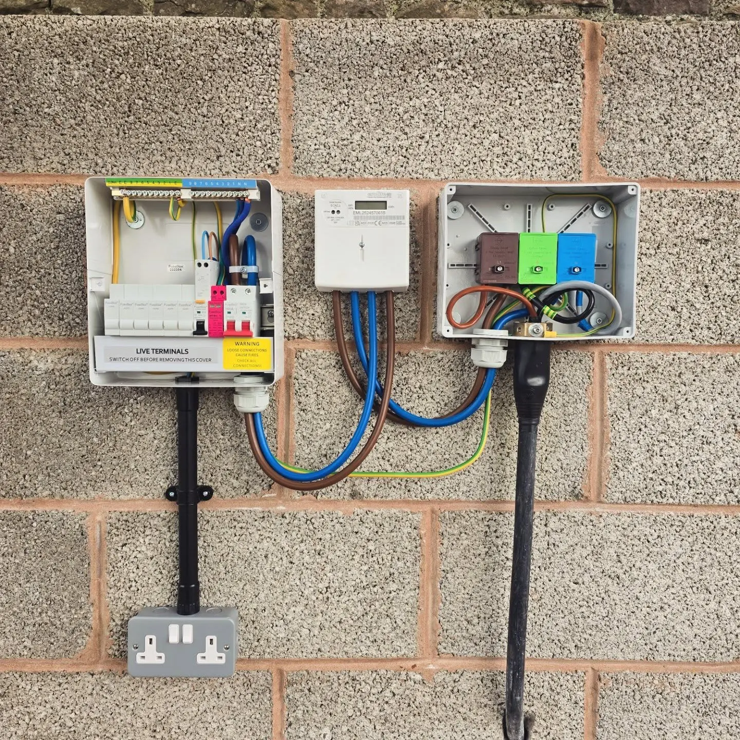 An electrical setup mounted on a cinder block wall, featuring an open consumer unit with circuit breakers, a digital electricity meter, and a second junction box with colored cable connectors, connected to a double wall outlet below.