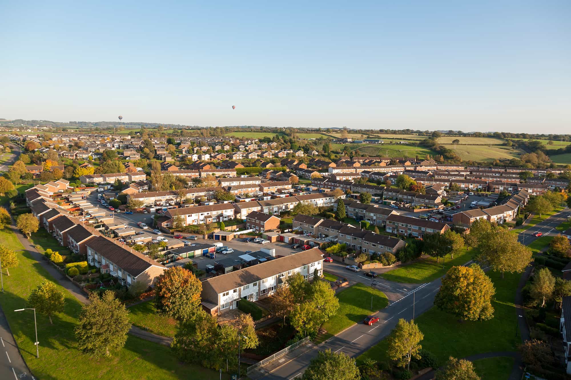 Aerial view of a residential neighborhood of terraced houses with green spaces, with two hot air balloons in the distance.