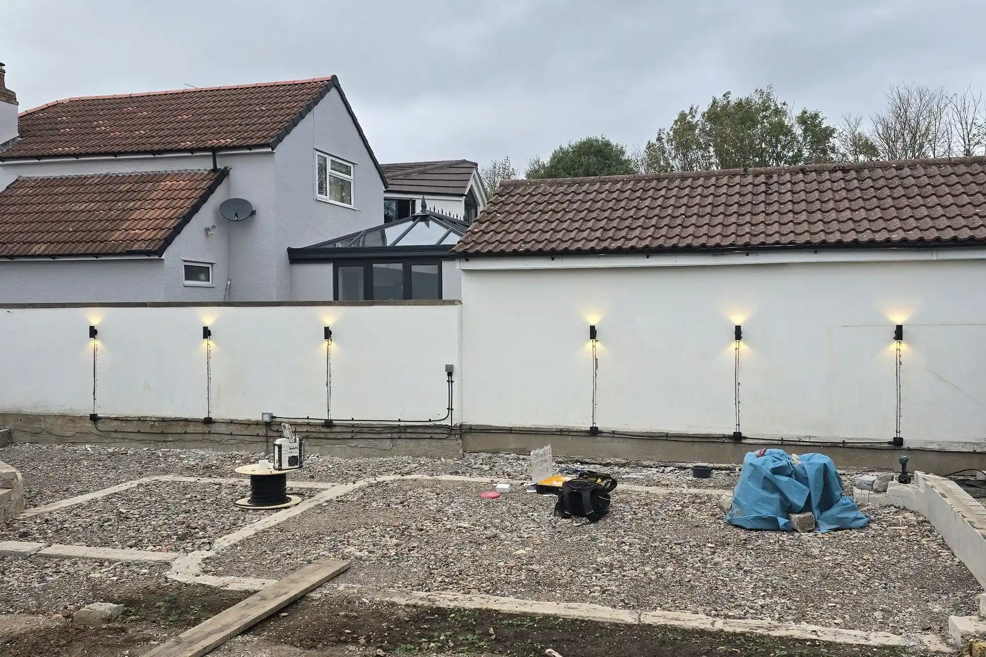 A backyard under construction featuring a white wall mounted with six glowing lights, situated in front of a house with a grey exterior and tiled roof.