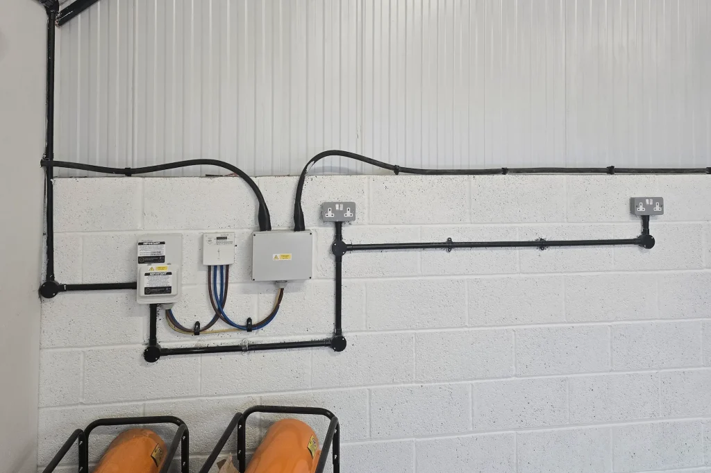 Electrical components and conduits mounted on a white brick wall, including an isolation switch, a consumer unit, a junction box, and two double electrical outlets.