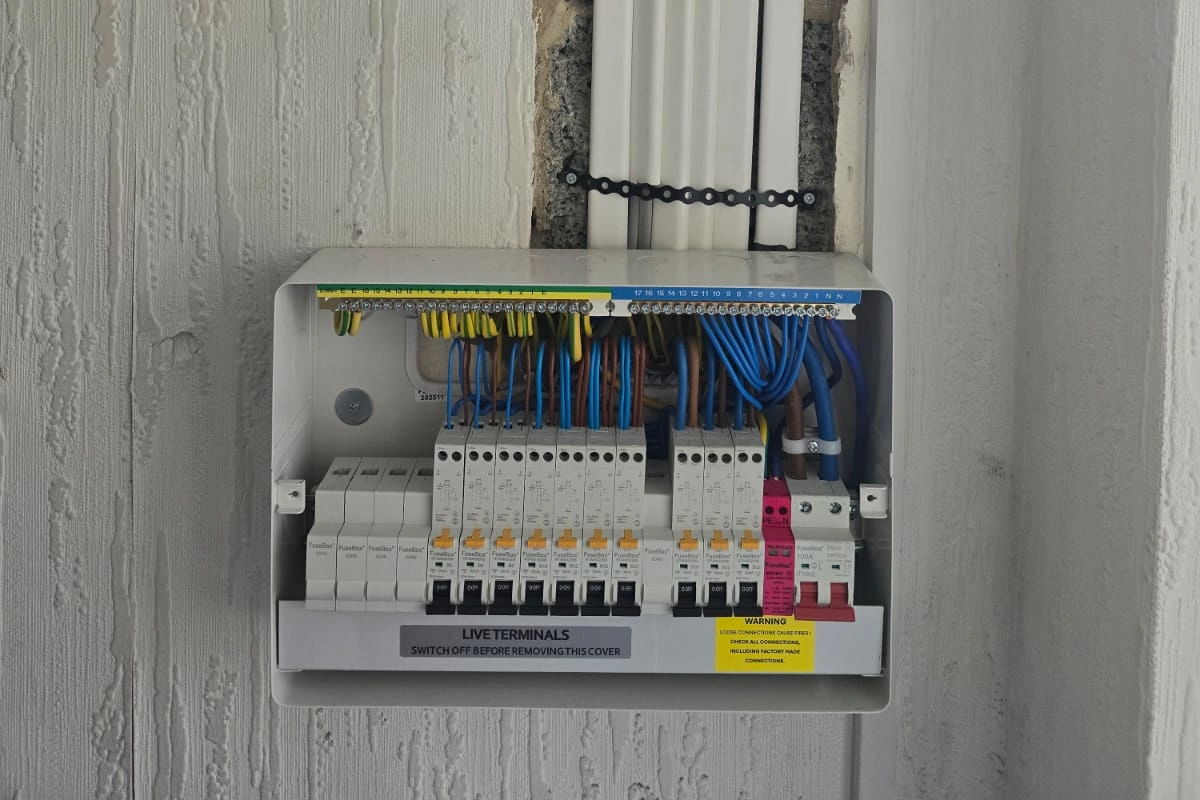 An open electrical consumer unit with exposed breakers, wiring, and warning labels mounted on a wall.